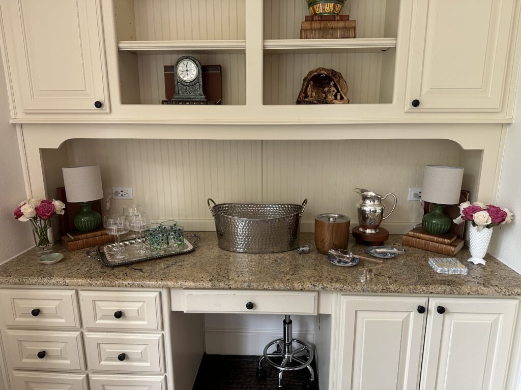 Home bar styled for entertaining with a silver serving tray, galvanized ice bucket, crystal glasses, small table lamps, and fresh pink and white roses