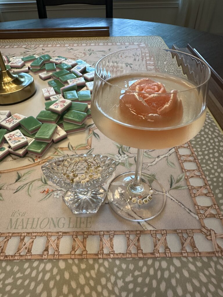Hugo Spritz cocktail in a crystal coupe with a rose shaped ice mold and personalized beaded wine charm on an It's A Mahjong Life Whispering Sparrow mat with Miss Mahjong Classic Jade tiles in the background