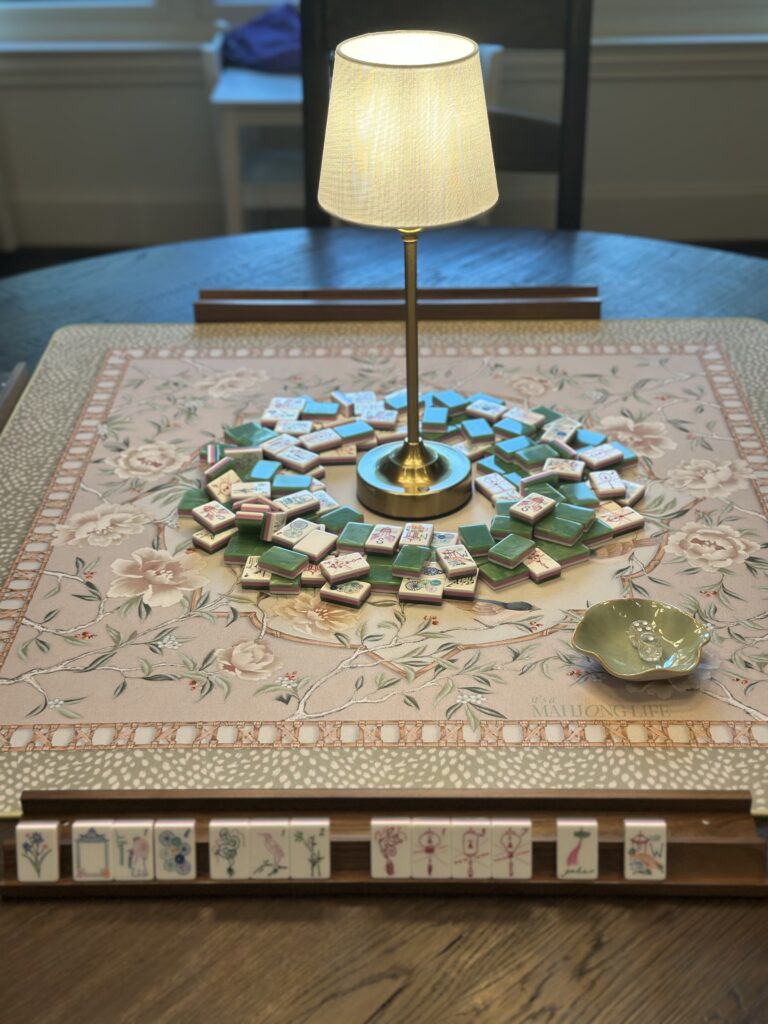 Miss Mahjong Classic Jade tiles arranged in a circle on an It's A Mahjong Life Whispering Sparrow mat with a brass cordless lamp and small celadon dish