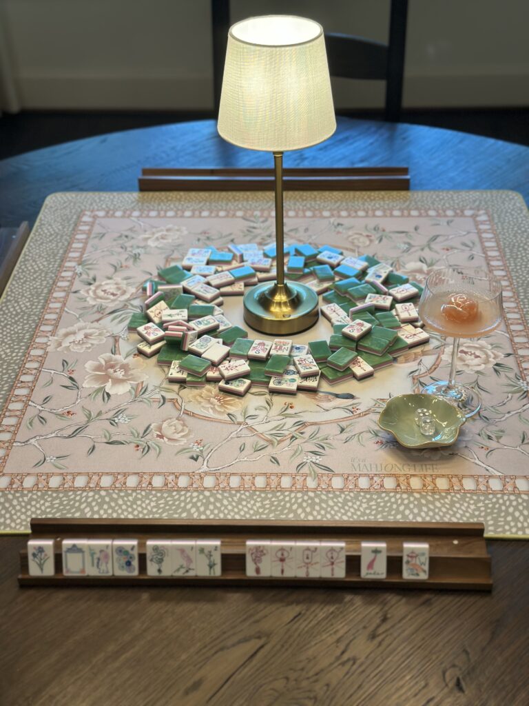 Miss Mahjong Classic Jade tiles arranged on an It's A Mahjong Life Whispering Sparrow mat with Bird Bam Boutique walnut racks, a Hugo Spritz cocktail with rose ice mold, and a brass cordless lamp