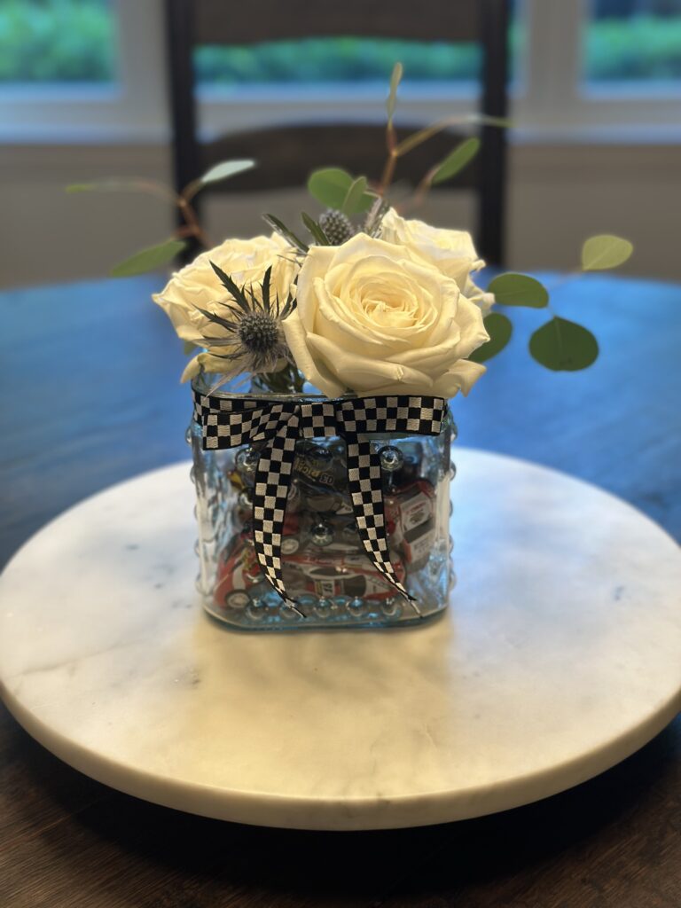 Glass hobnail vase with checkered ribbon holding white roses and toy cars.
