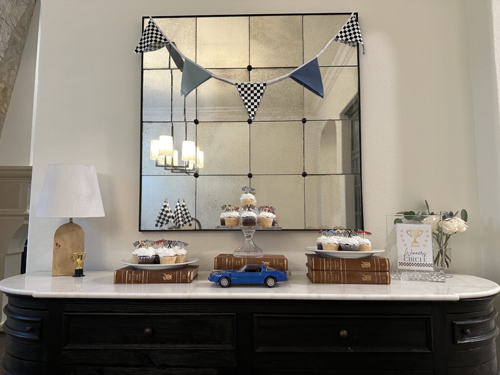 Cupcakes and a crystal cake stand on a sideboard beneath a checkered bunting mirror.