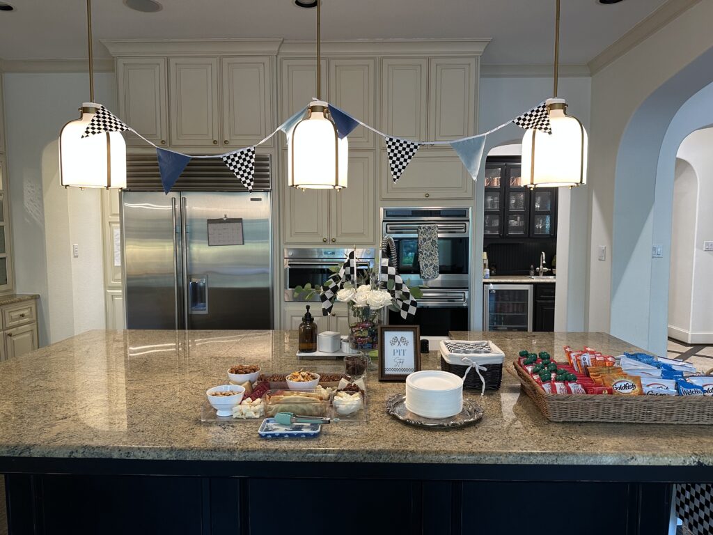 Kitchen island snack spread with cheese board, pretzels, and Goldfish under a checkered pennant garland.