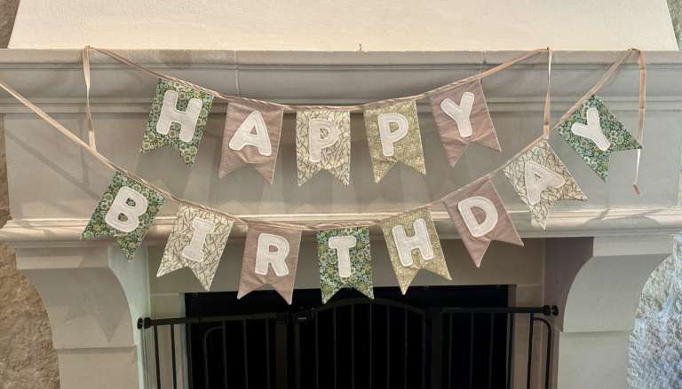 A 'HAPPY BIRTHDAY' fabric bunting banner in sage green floral and cream Tilda Fabrics prints with a pink striped pennant, hung on a stone fireplace mantel.